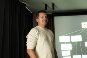 Uśmiechnięty mężczyzna w jasnym swetrze przemawia przed publicznością